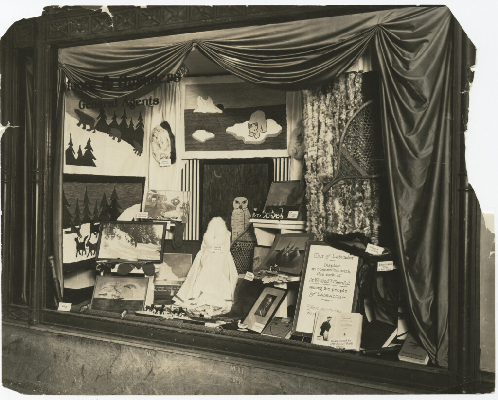 Display of goods in window of "Moore & Summers General Agents." Items on display include books by Wilfred T. Grenfell, photographs, whips, fur hats, hooked rugs and snowshoes.